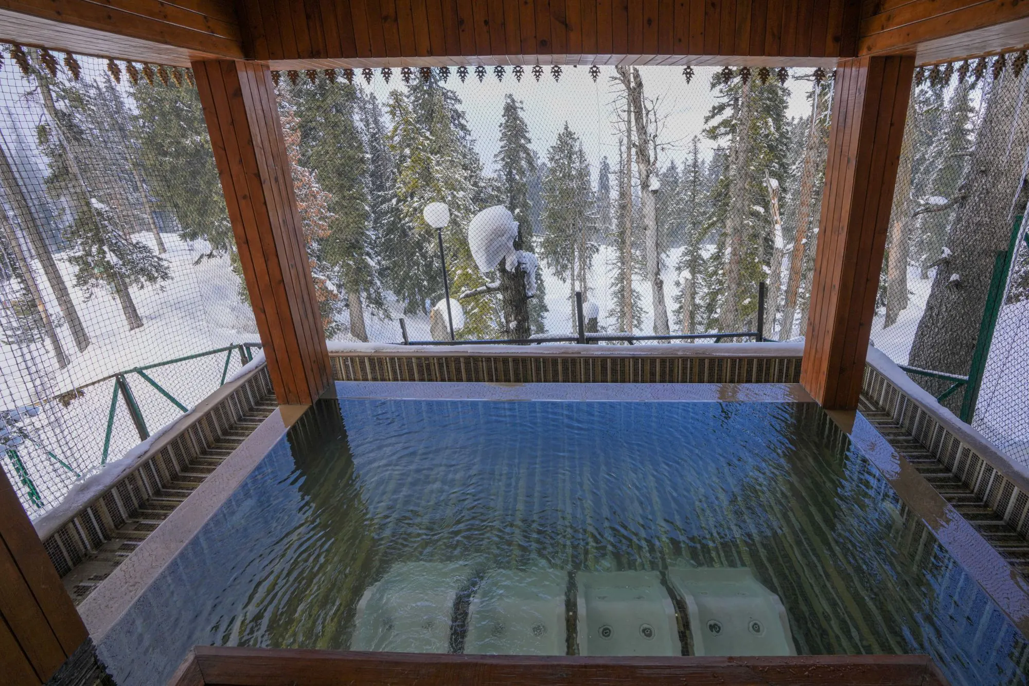 Open-sided pool pavilion with warm water facing a snow-covered pine forest
