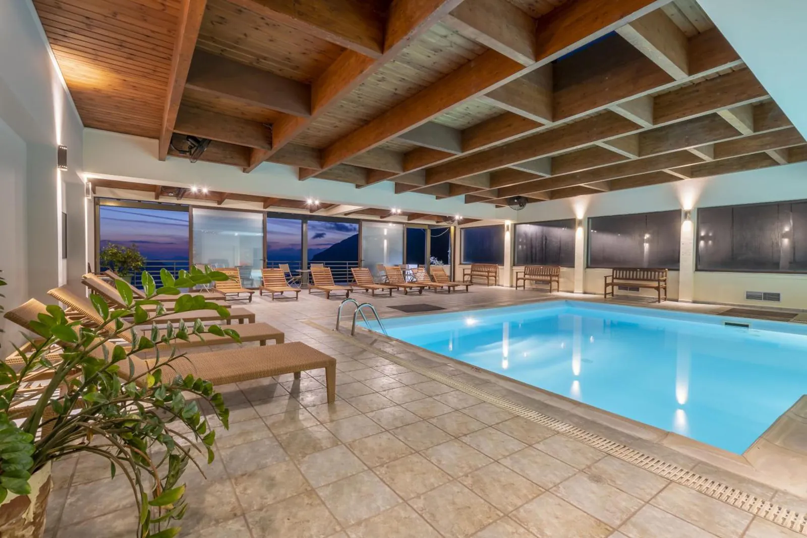Indoor pool and sun loungers under a timber ceiling with floor-to-ceiling windows at dusk