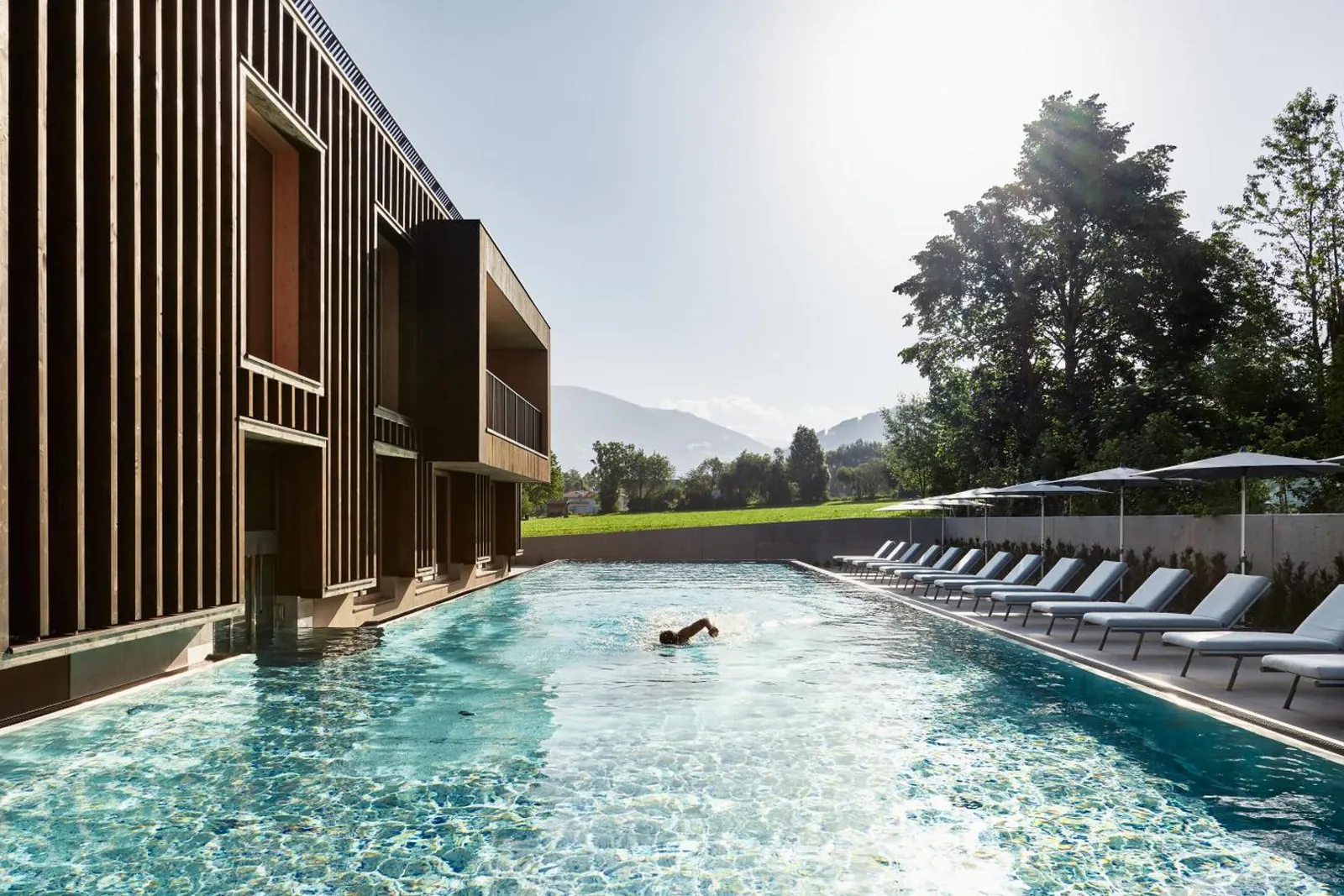 Swimmer doing lengths in the 25-metre sports pool beside the timber facade, loungers along the meadow side
