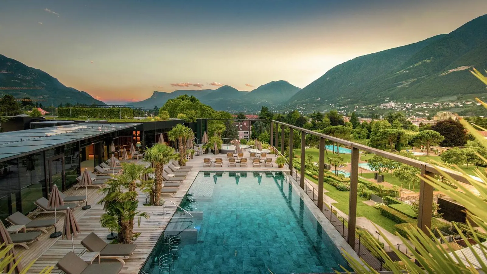 Rooftop pool lined with palms and loungers at dusk, mountains behind