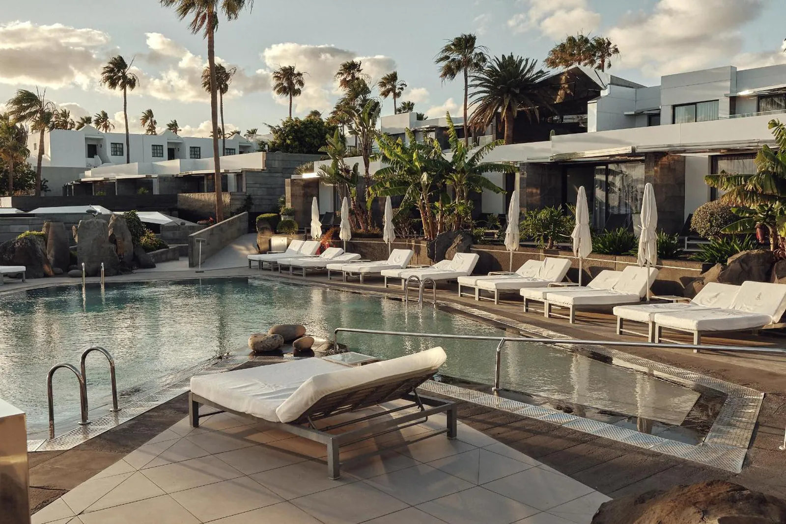 Row of white loungers along the pool with low white suites and palms behind