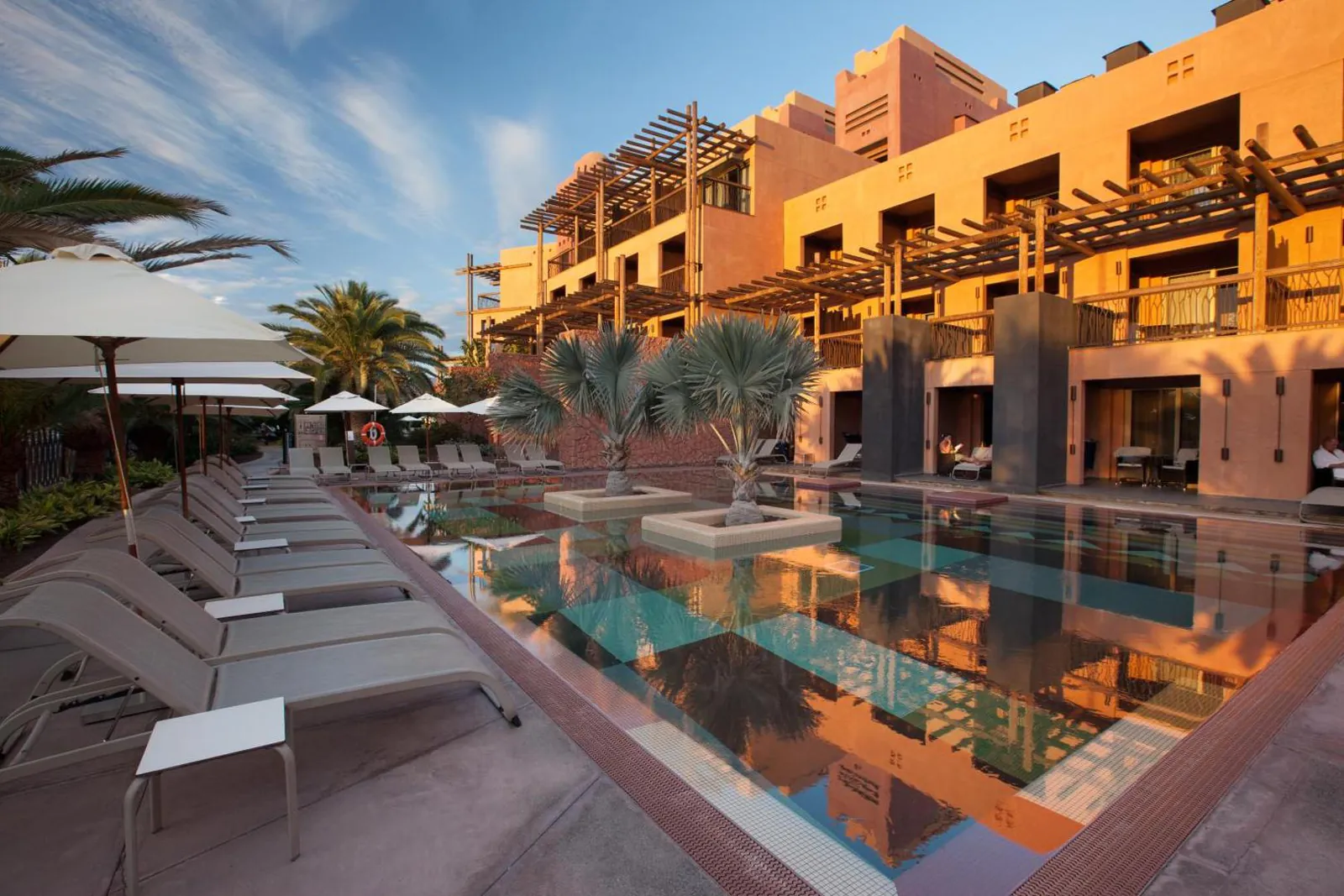 Long pool running past the terracotta buildings, with loungers along its edge