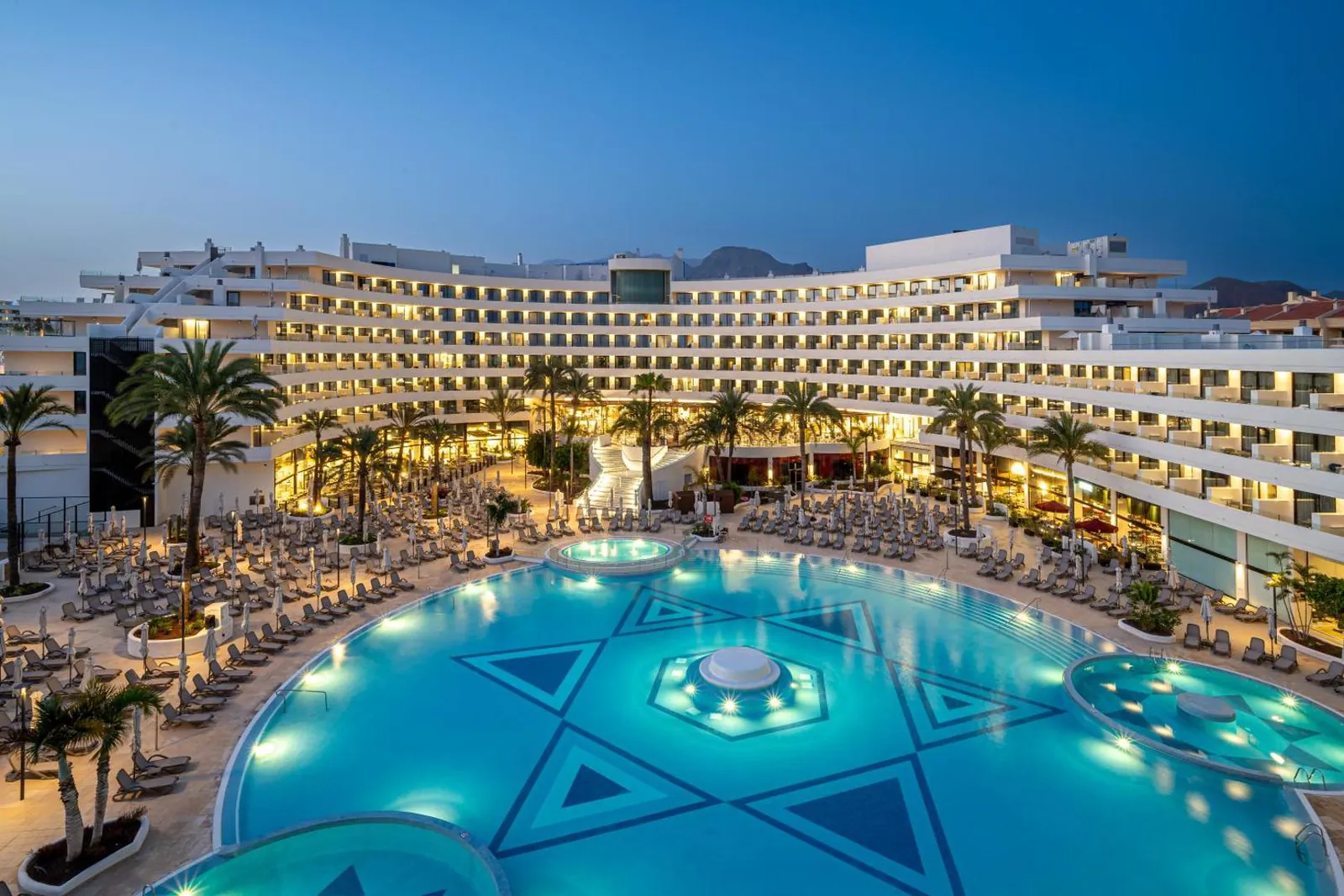The large pool lit up at dusk with its patterned tiled floor and the curved hotel behind