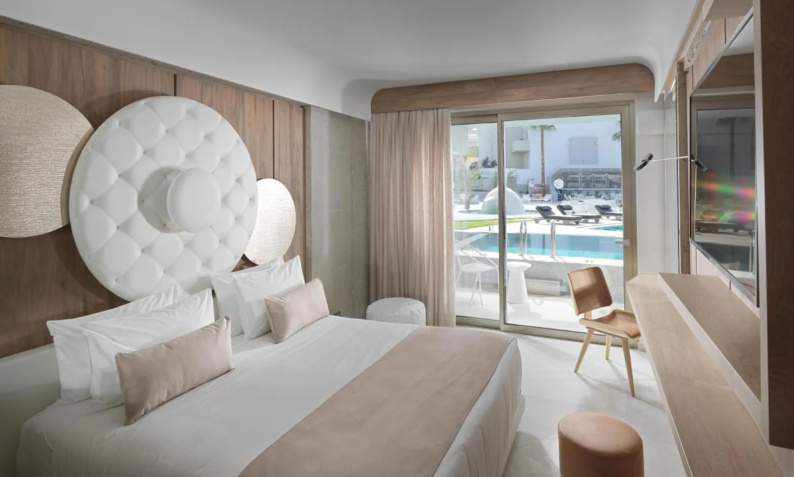Bedroom with a round padded headboard and glass doors onto a pool-view balcony