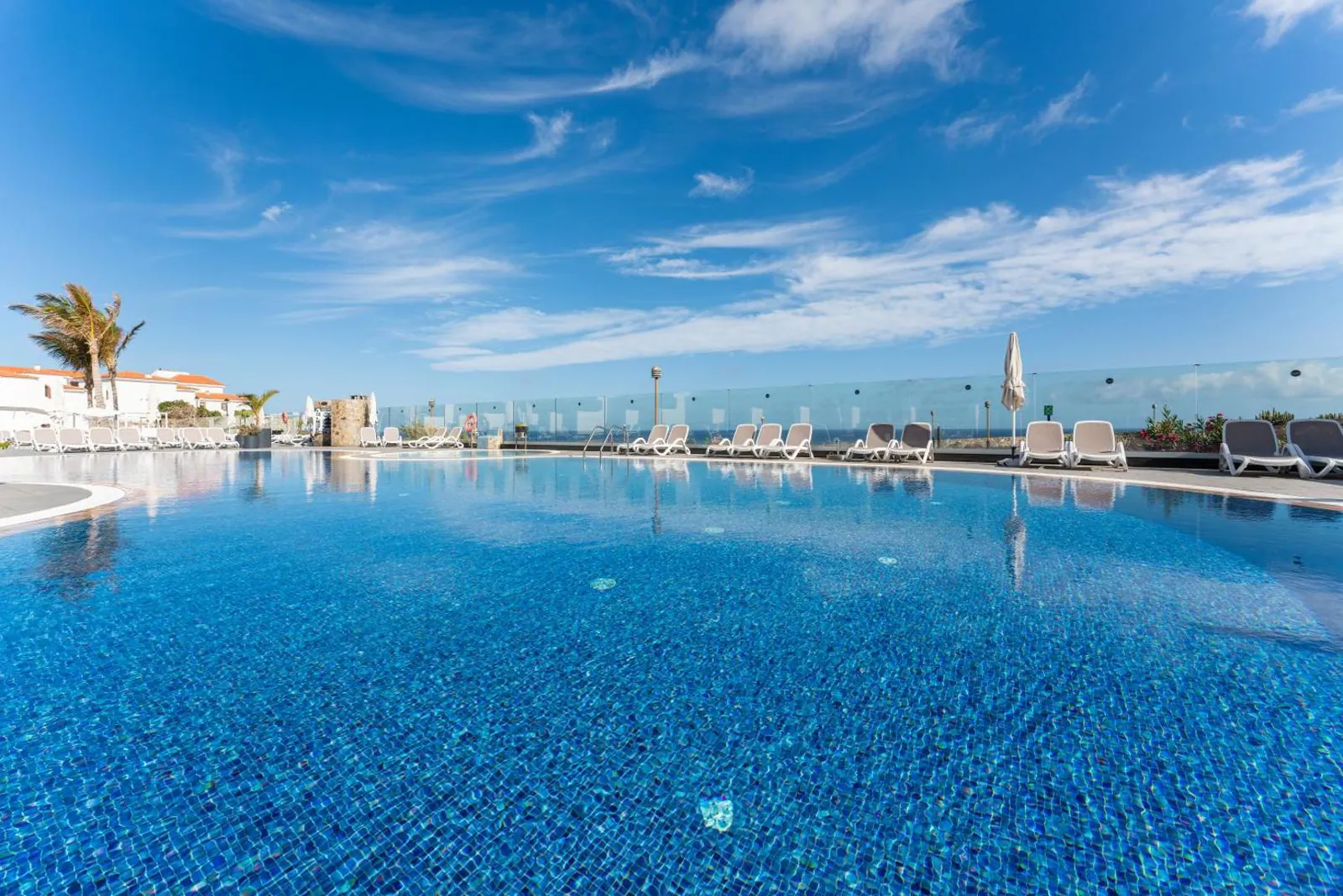 Outdoor pool lined with loungers under a blue sky, the sea just past the glass railing