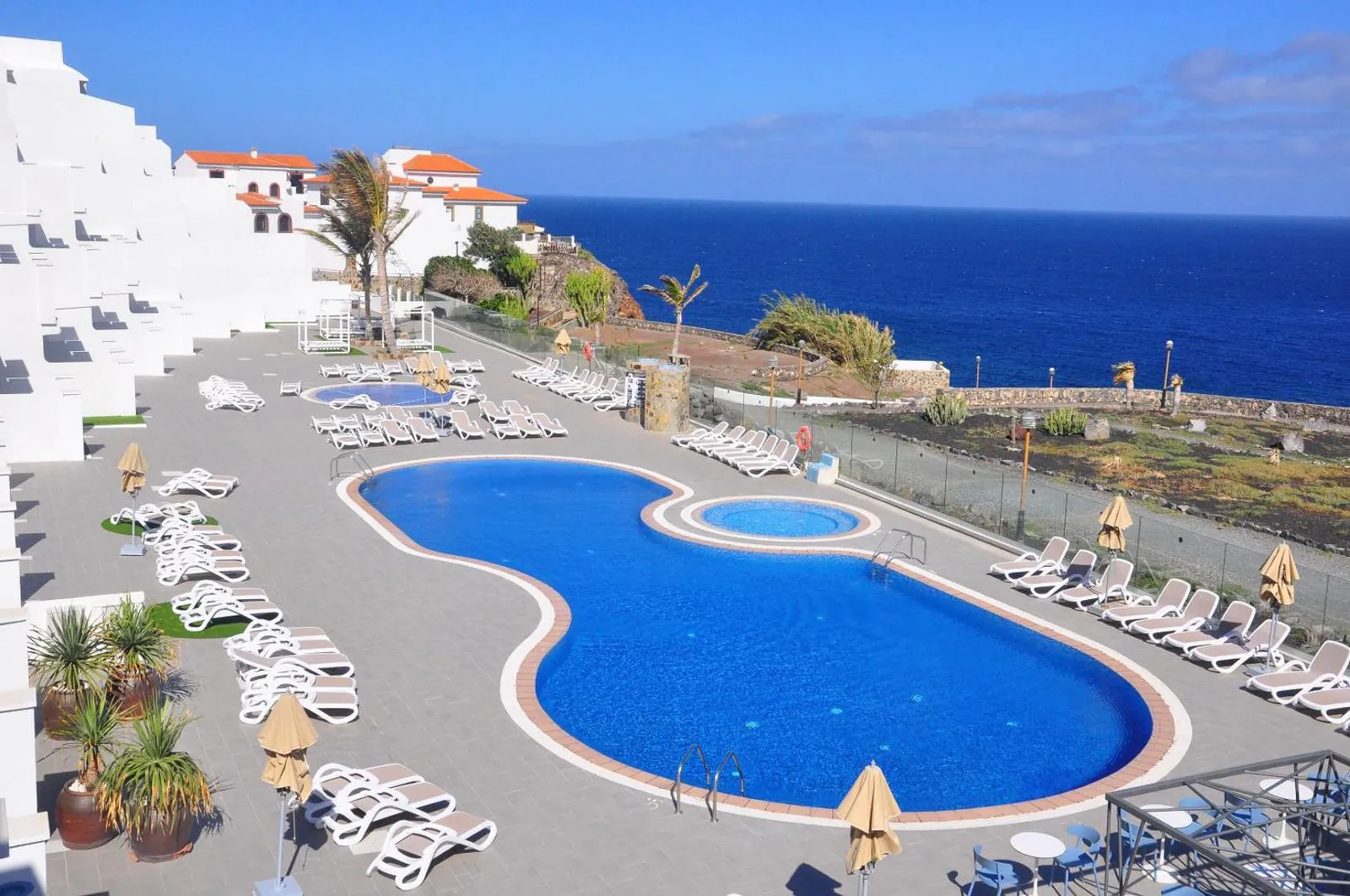 Kidney-shaped pool with a round hot tub and rows of loungers above the sea