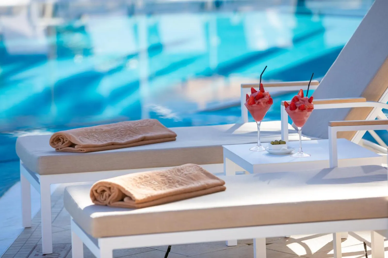 Two poolside loungers with towels and drinks beside turquoise water